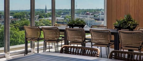 Un área de comedor exterior moderna con sillas de mimbre y una mesa, ventanas amplias con vistas a la ciudad y un televisor de pantalla plana montado en la pared.