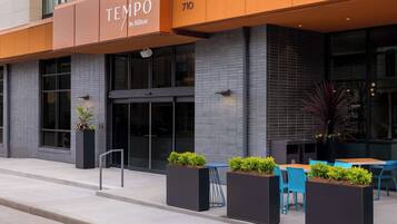 L’extérieur d’un hôtel au design moderne, avec une enseigne indiquant « TEMPO by Hilton » et l’adresse « 710 ». On y trouve des espaces extérieurs aménagés avec des tables et des chaises, ainsi que des jardinières remplies de verdure.