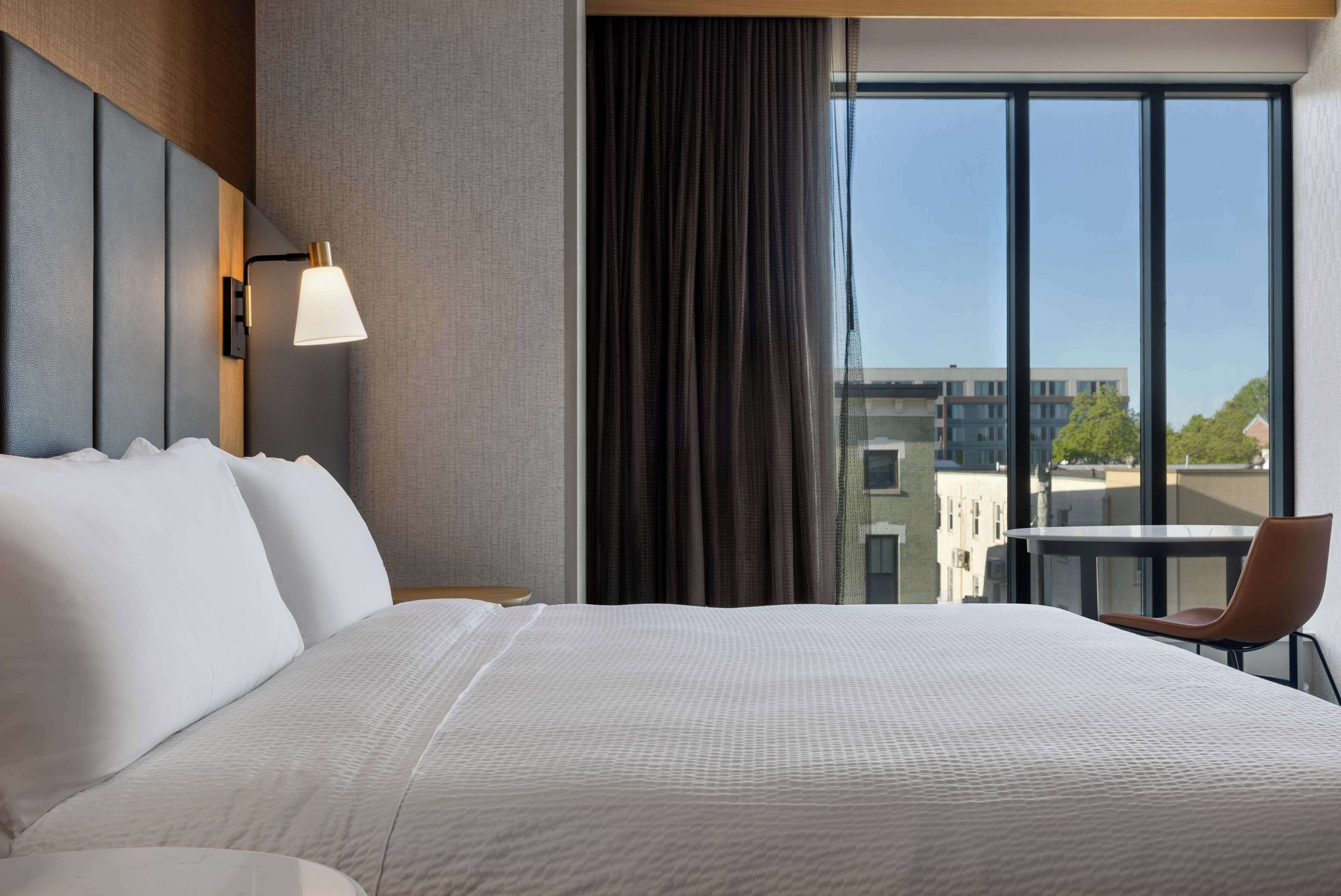 A hotel room with a large bed, a desk with a chair, a window with a view of buildings, and a wall-mounted lamp.
