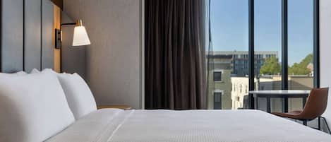 A hotel room with a large bed, a desk with a chair, a window with a view of buildings, and a wall-mounted lamp.