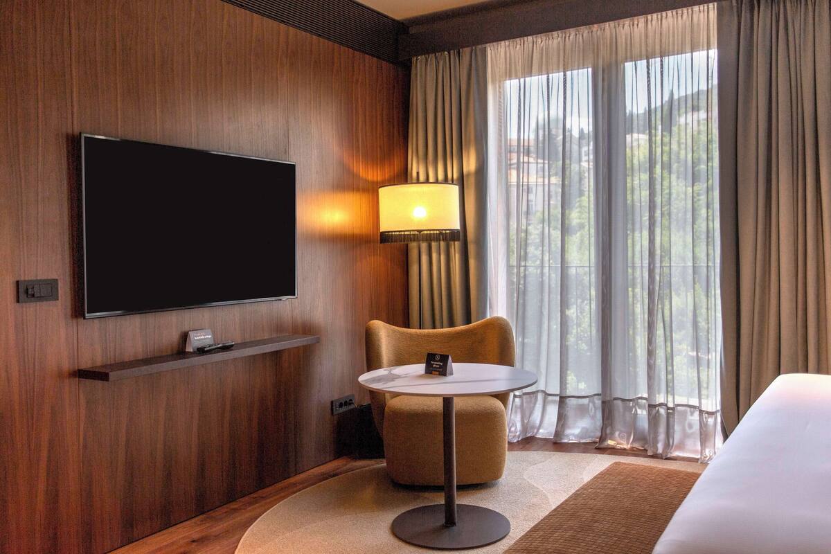 Une chambre d’hôtel dotée d’un téléviseur à écran plat fixé au mur, d’une petite table avec une lampe, d’un fauteuil confortable et d’une grande fenêtre avec des rideaux.