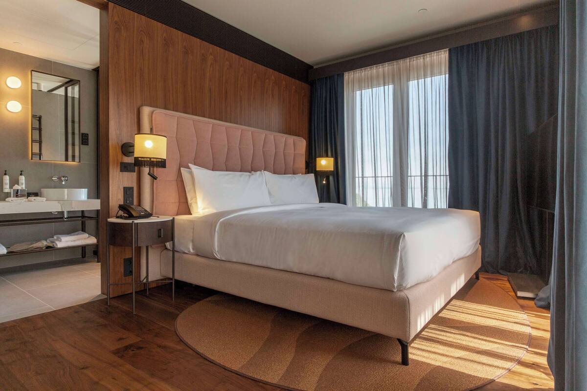 Une chambre d’hôtel moderne avec un grand lit, un mur décoratif en bois, un tapis rond, une table de chevet avec une lampe, une coiffeuse avec un miroir et une grande fenêtre avec des rideaux.