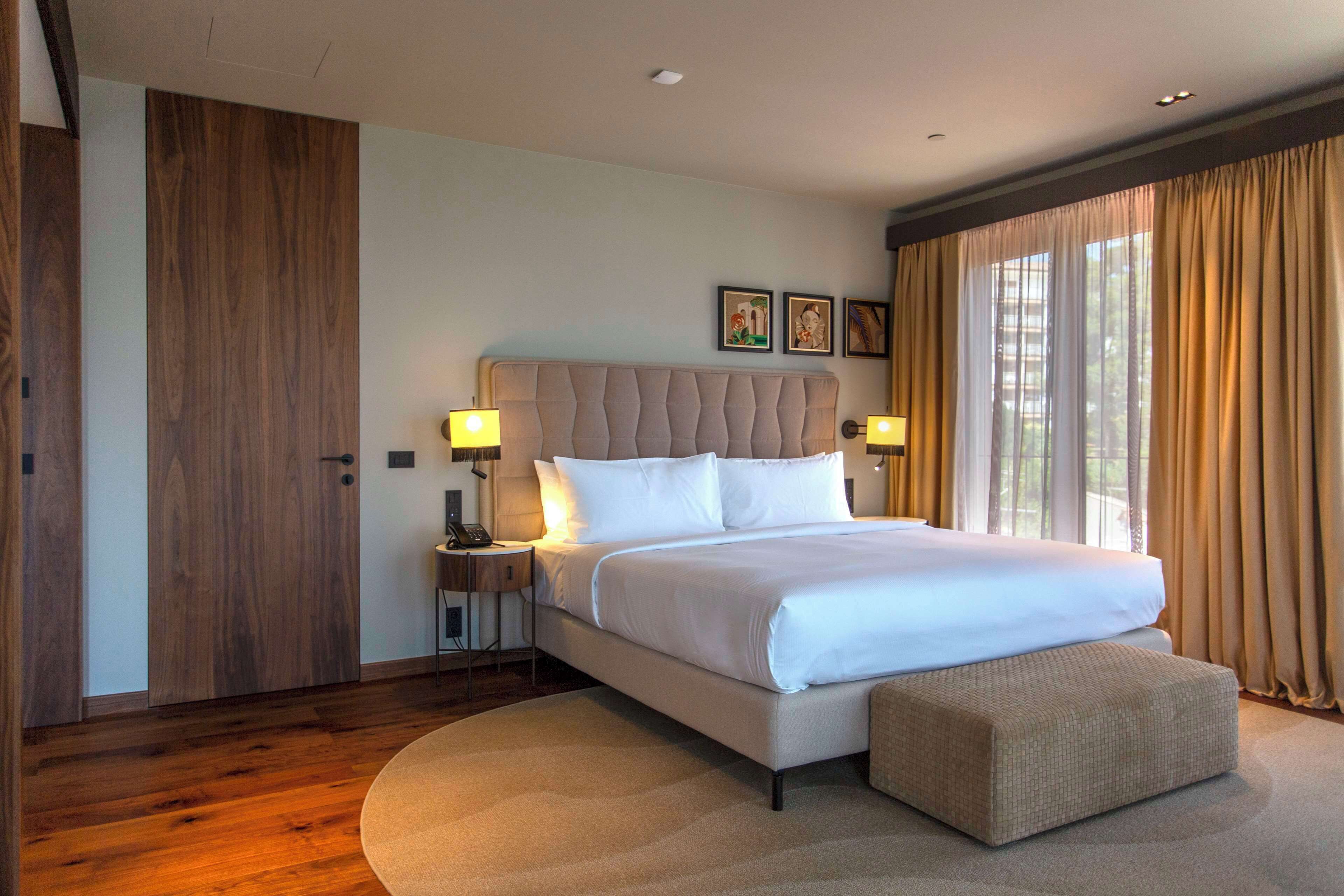 A modern hotel room with a large bed, bedside lamps, a wooden wardrobe, and a large window with curtains.