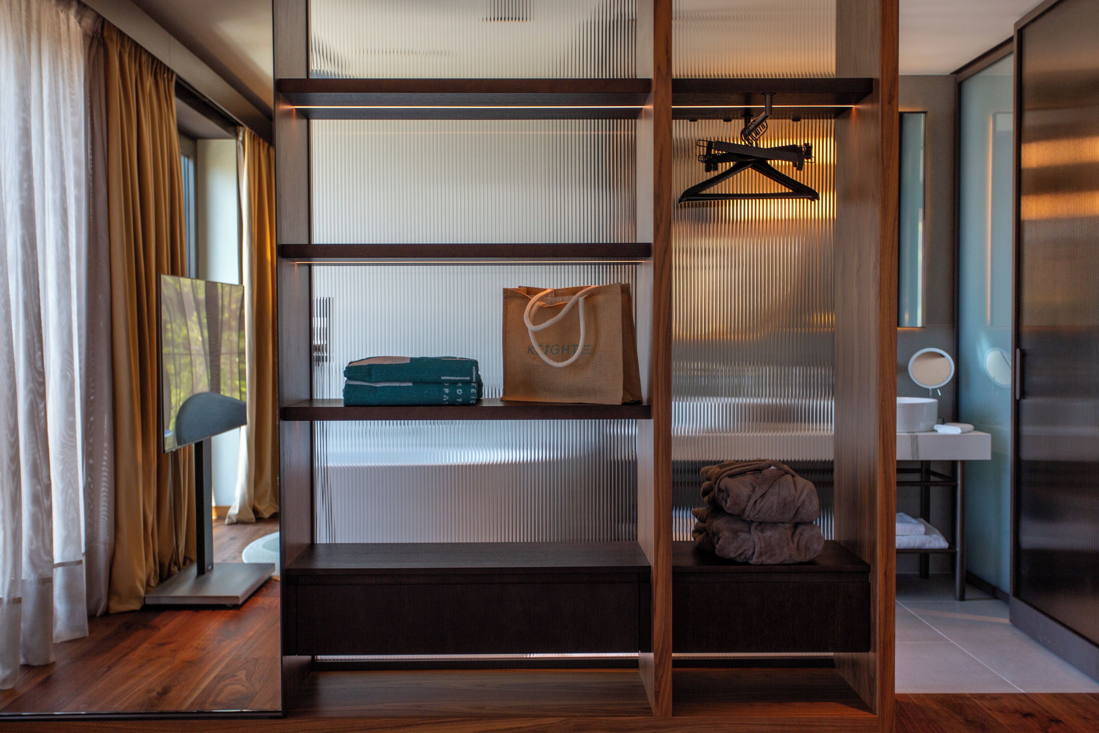 A modern hotel room with a wooden floor, a built-in shelf unit with a hanging garment rack, a small seating area, and a bathroom with a mirror and sink.