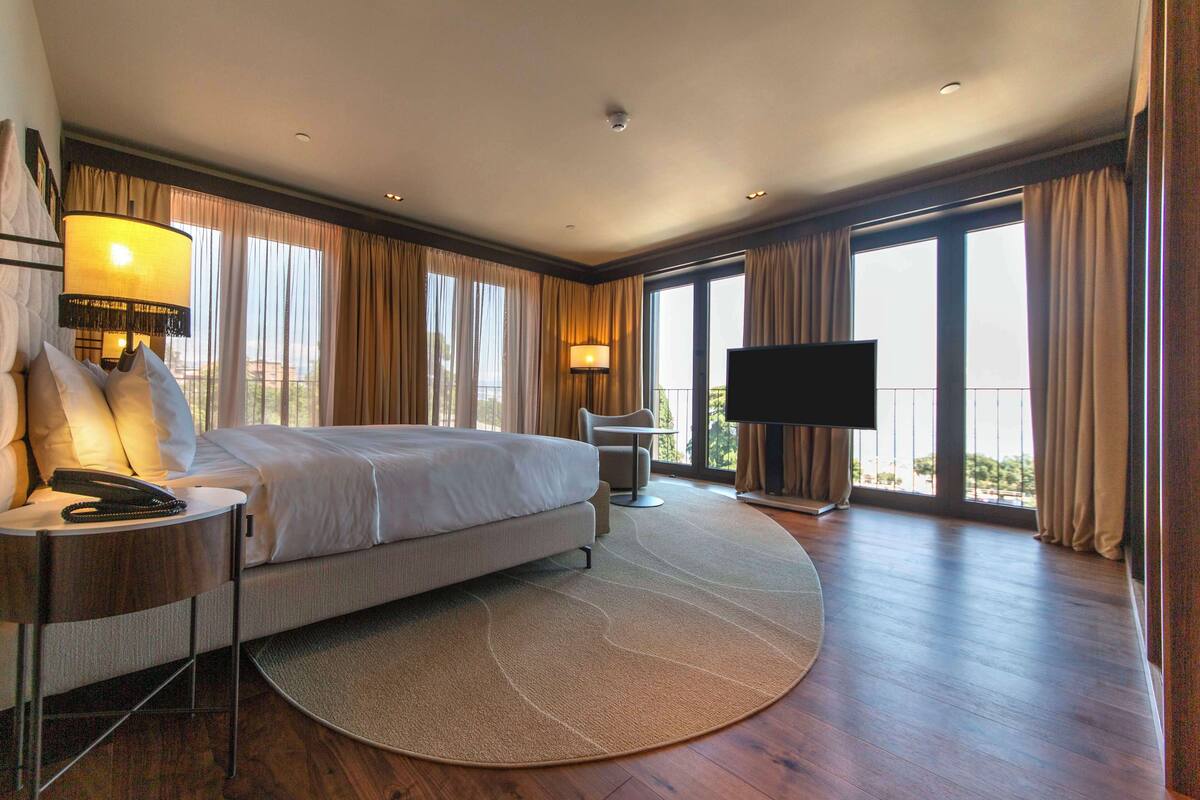 Une chambre d’hôtel spacieuse, dotée d’un grand lit, d’un bureau avec une lampe, d’une télévision et d’un balcon avec des rideaux.