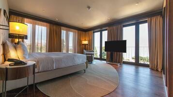 A spacious hotel room with a large bed, a desk with a lamp, a TV, and a balcony with curtains.