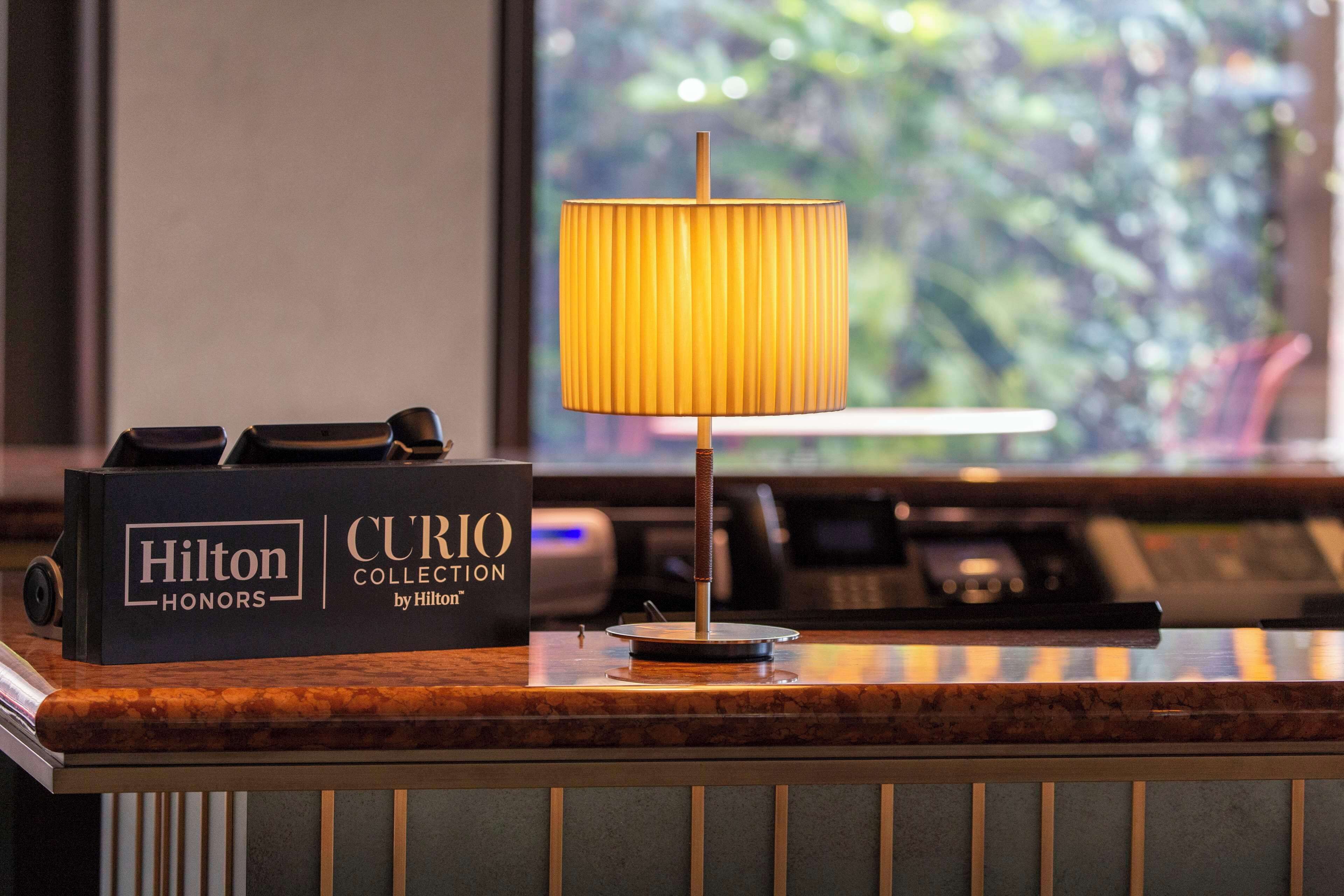 Un mostrador de recepción de hotel con un letrero de Hilton Honors, una lámpara y vistas al verdor a través de la ventana.