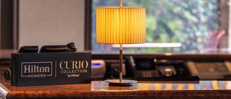A hotel reception desk with a Hilton Honors sign, a lamp, and a view of greenery through the window.