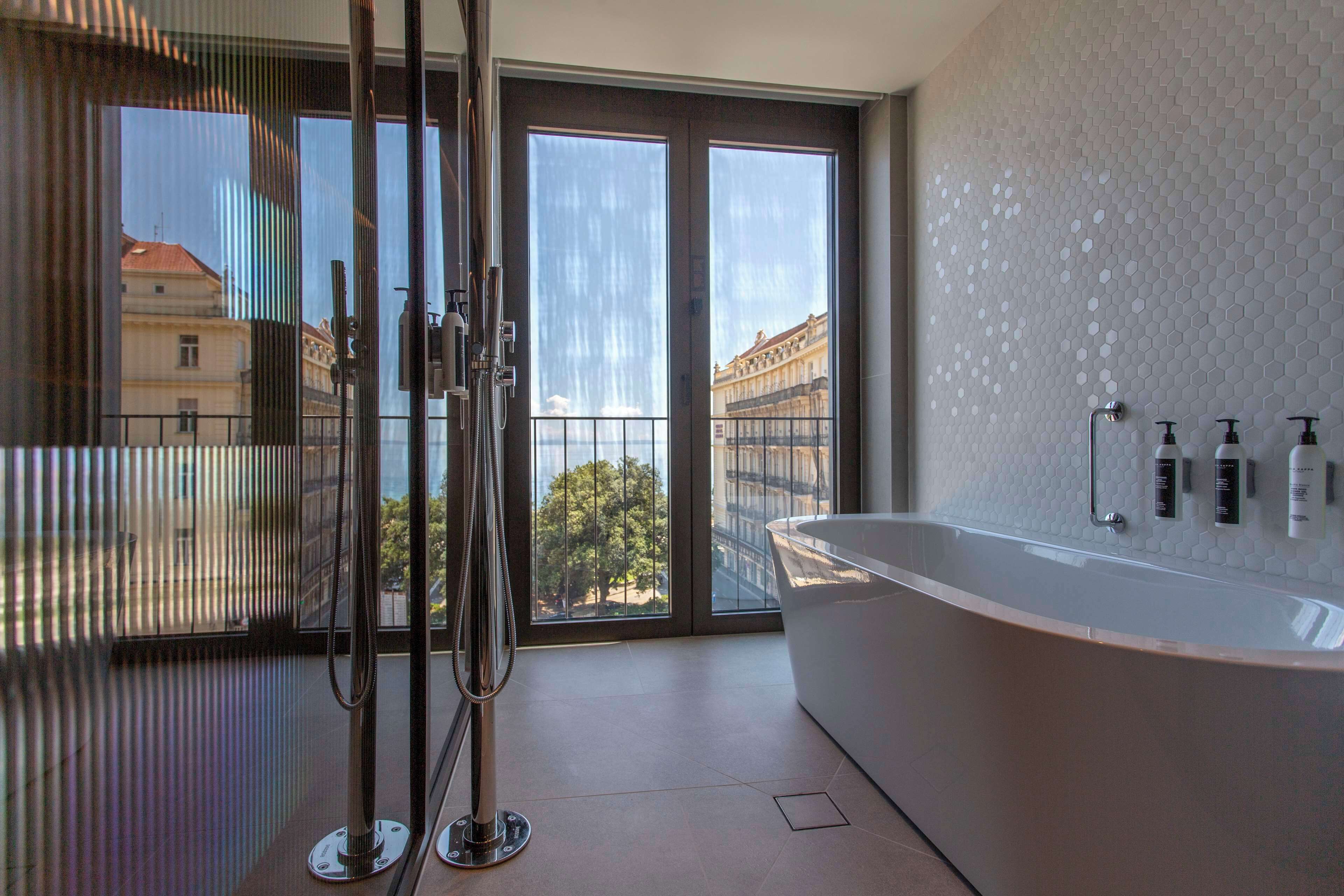 Un baño moderno con una bañera grande, cabina de ducha de vidrio y vistas a edificios a través de amplios ventanales.