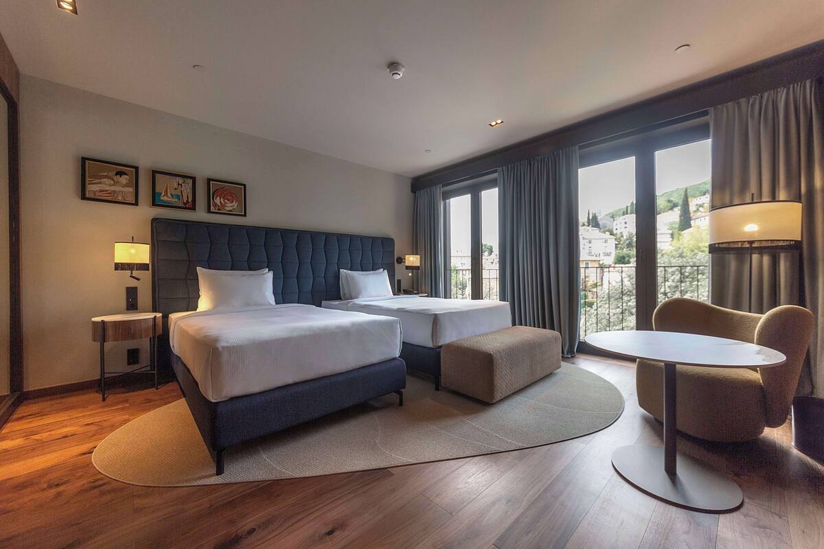 Une chambre d’hôtel avec un grand lit, deux tables de chevet, une table ronde et une vue sur la ville grâce à de grandes fenêtres.