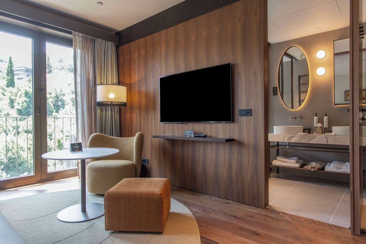 Une chambre d’hôtel moderne dotée d’une grande télévision, d’un canapé, d’une petite table, d’un pouf rond, d’une salle de bain avec un miroir et offrant une vue sur l’extérieur.