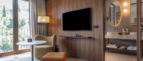 A modern hotel room with a large TV, a sofa, a small table, a round ottoman, a bathroom with a mirror, and a view of the outdoors.