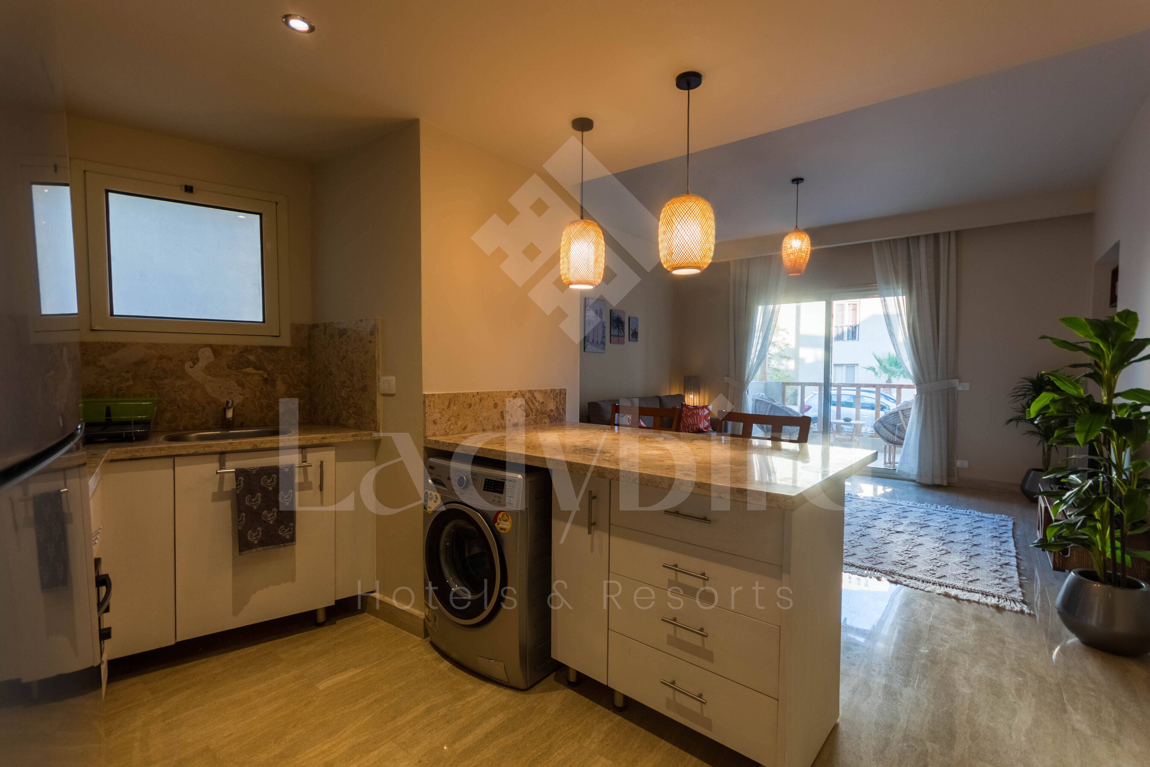 Una cocina moderna con lavadora integrada, una isla grande con pileta, luces colgantes y vistas a un balcón.