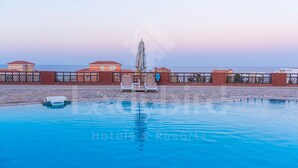 Una piscina con sillones de baño y sombrillas, rodeada por una valla y edificios con techos rojos.