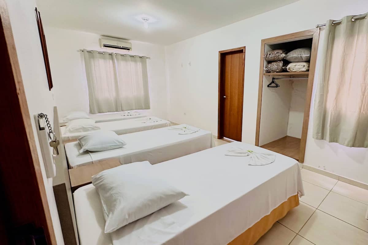 Habitación de hotel con dos camas individuales, un armario de madera, un baño con cortina de ducha y un armario pequeño con estantes.