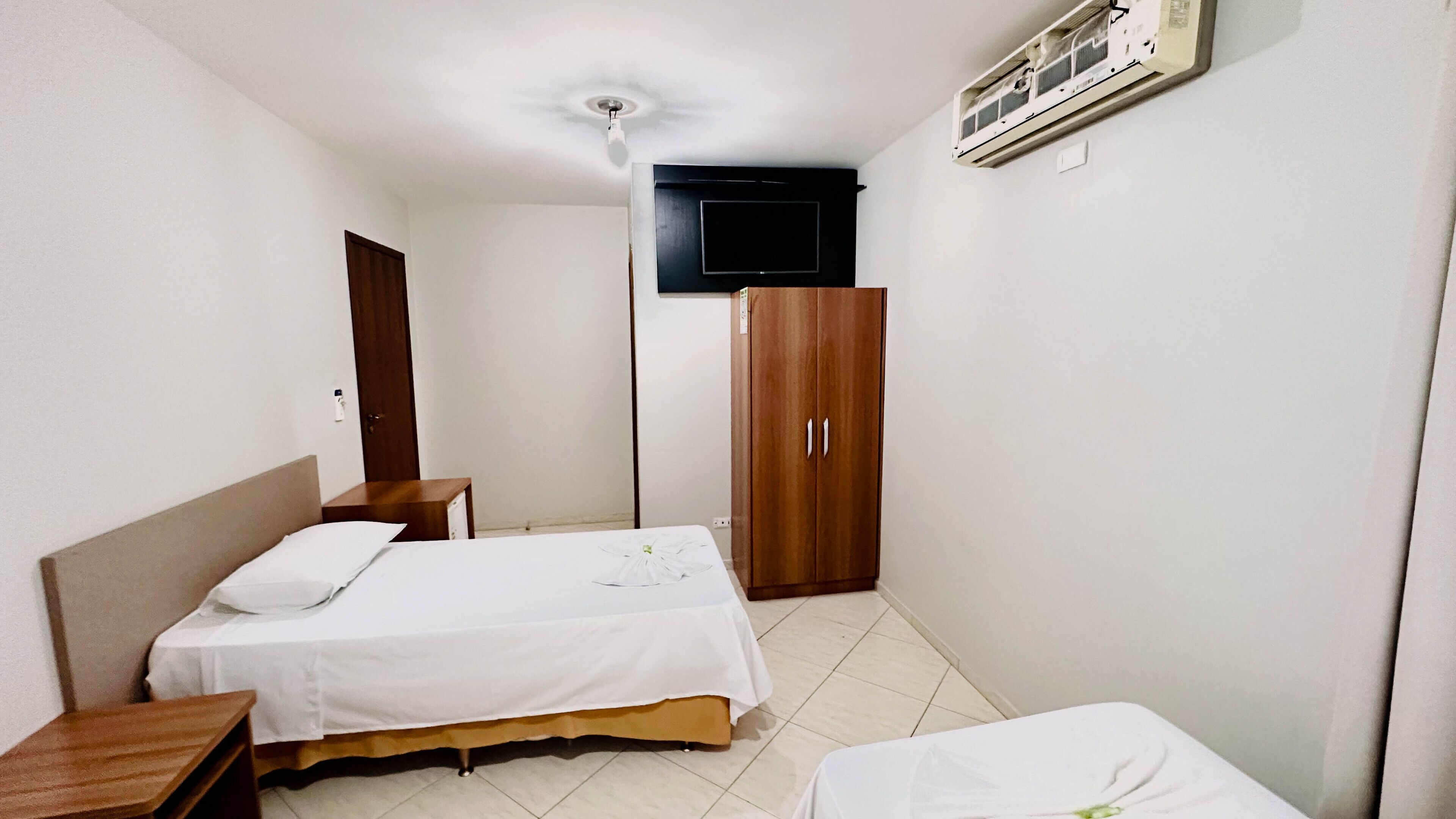 A small, clean room with a bed, a wooden wardrobe, a desk, and a television mounted on the wall.
