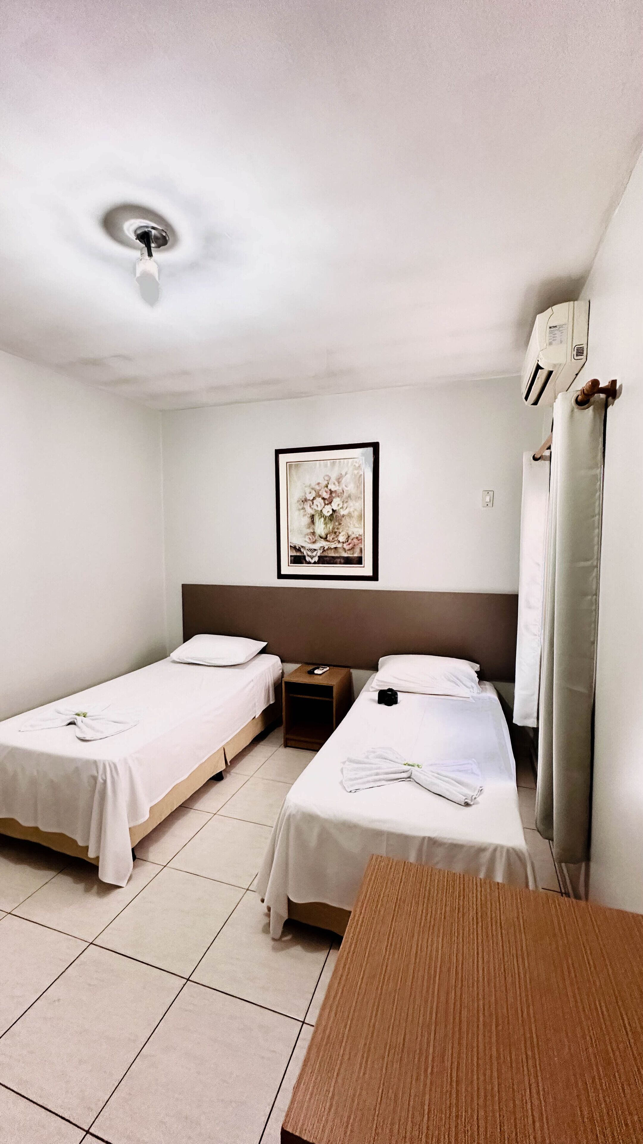 A hotel room with two single beds, a wooden table, a framed picture on the wall, and a ceiling fan.