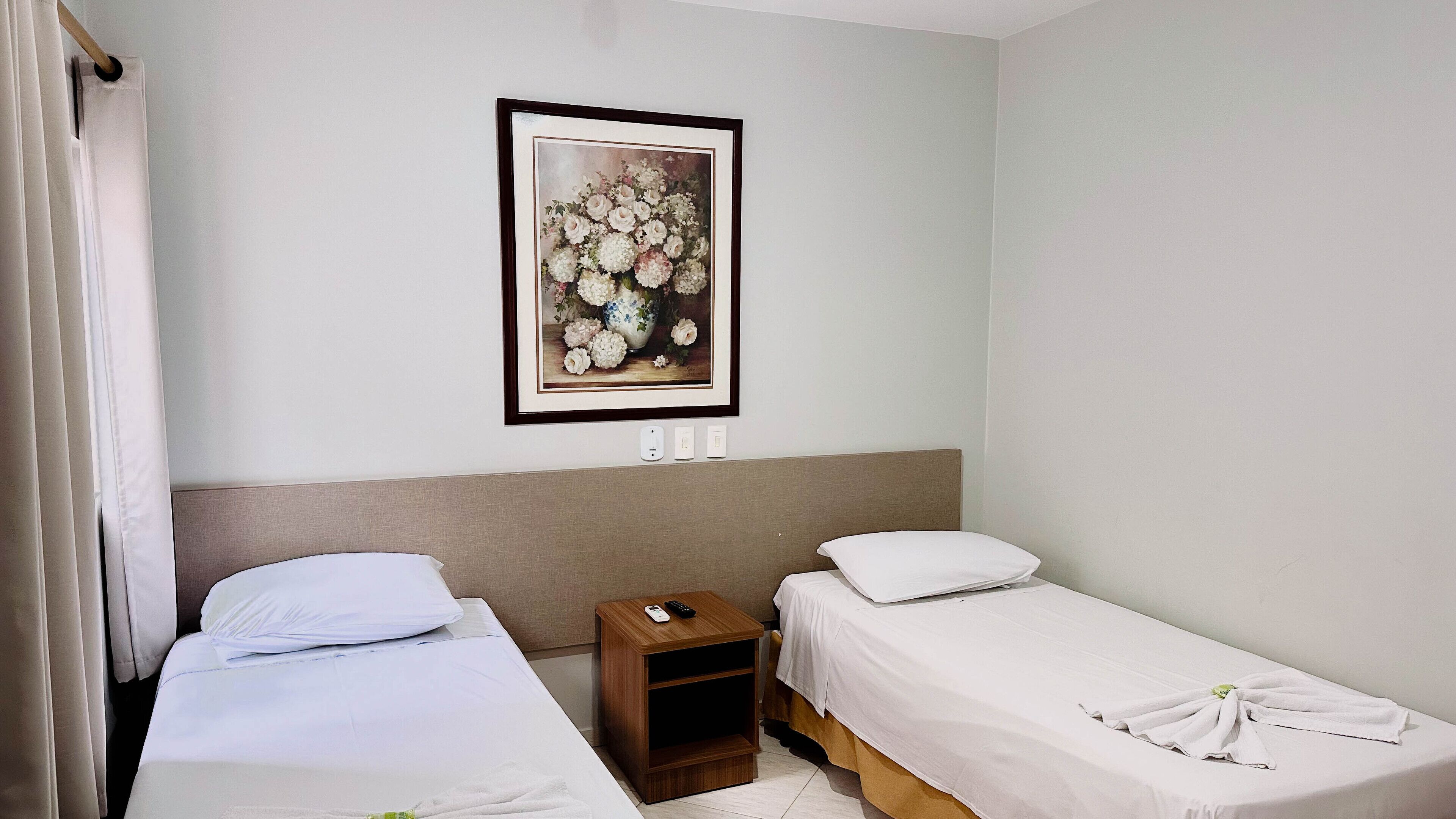 A hotel room with two single beds, a nightstand with a remote control, and a framed floral artwork on the wall.