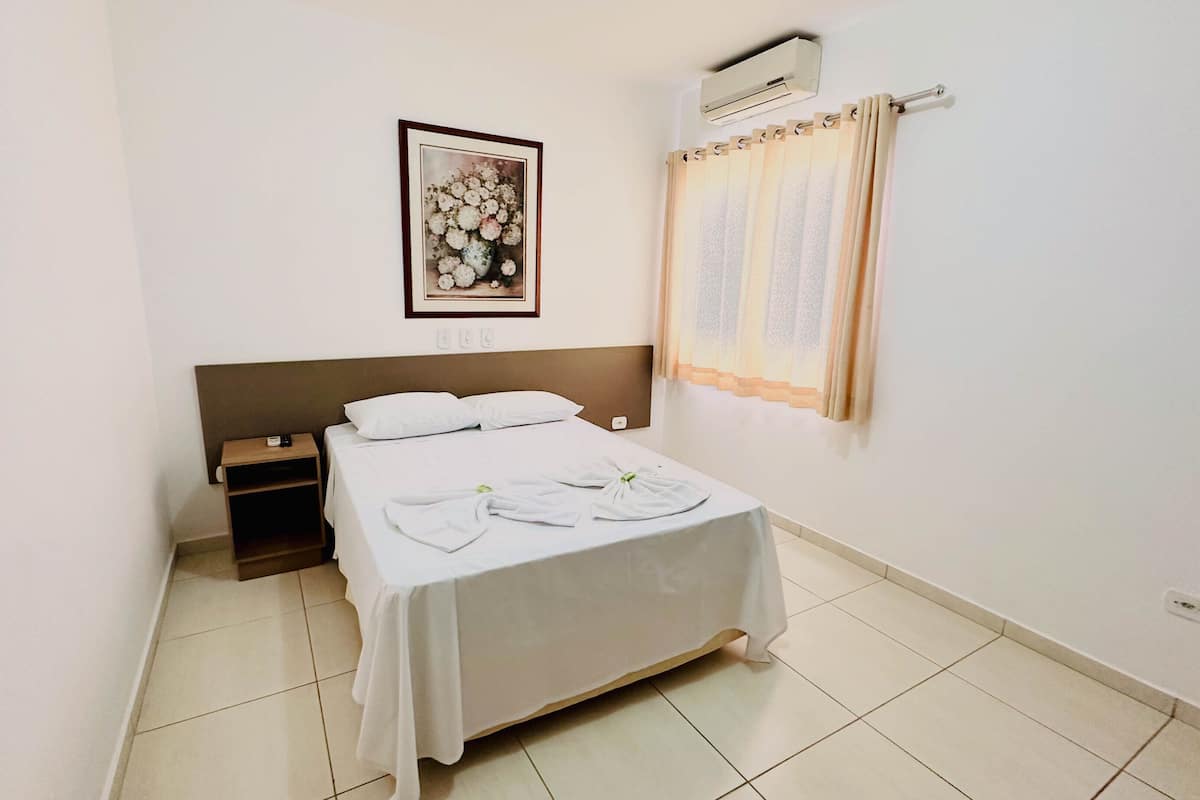 Una habitación de hotel con cama, mesita de noche, un cuadro en la pared, unidad de aire acondicionado y ventana con cortinas.