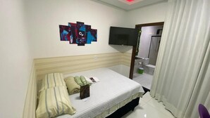 Une chambre à coucher avec un lit, un téléviseur à écran plat fixé au mur et une salle de bain visible à travers une porte ouverte.