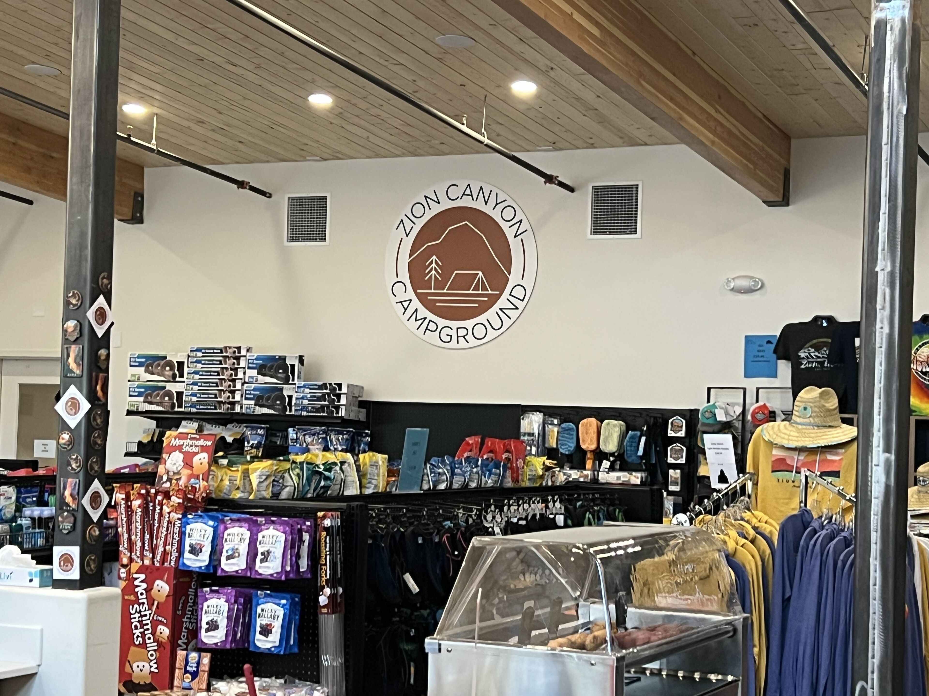 Een winkel met een 'Campground' bord, schappen vol snacks en een kledingrek.