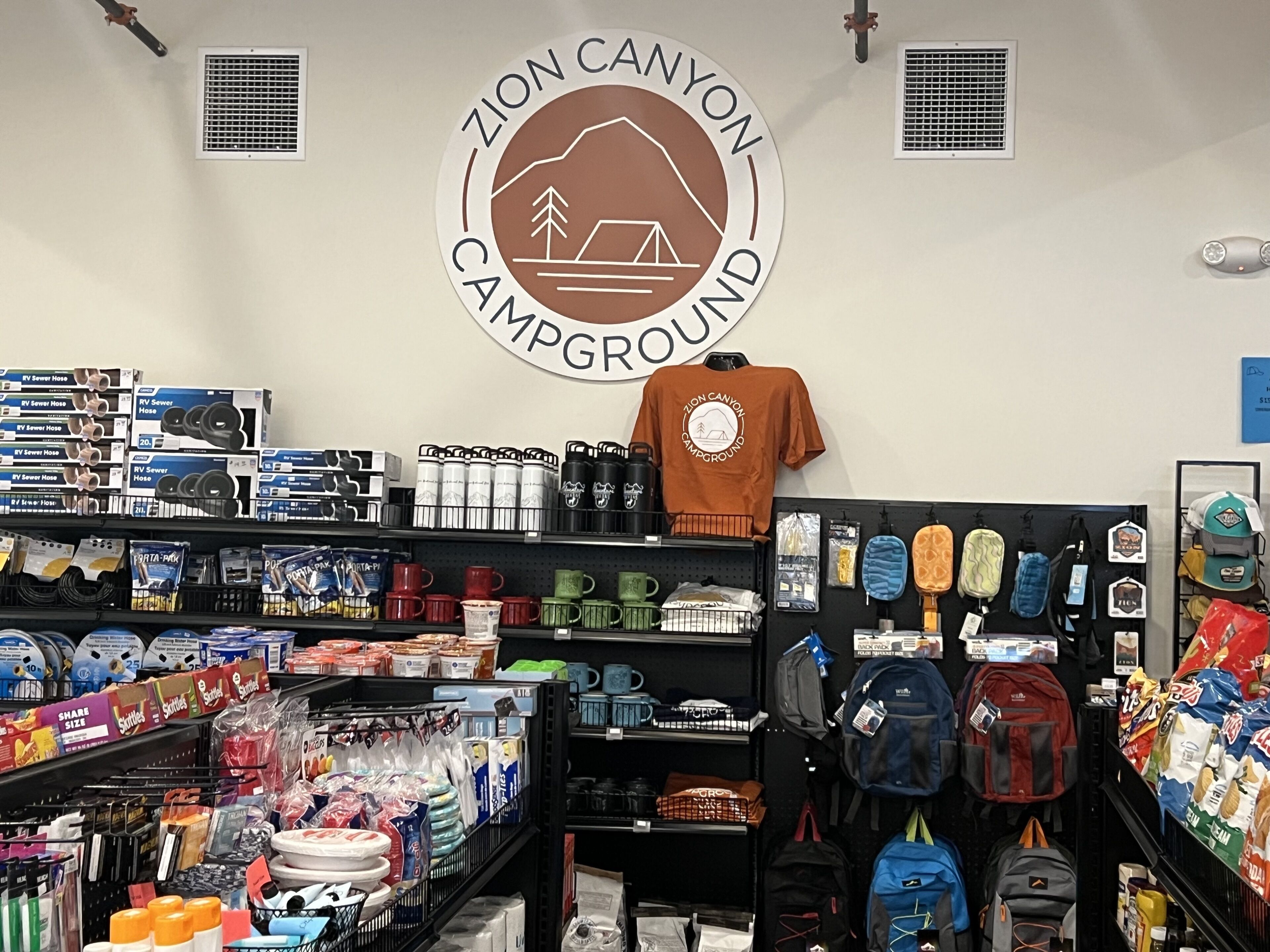 Loja com prateleiras repletas de equipamentos de camping, incluindo mangueiras para despejo de trailers, garrafas de água e mochilas.