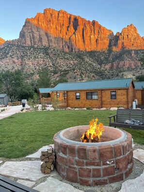 A rustic cabin with a fire pit in the foreground, surrounded by a well-maintained lawn and a mountain backdrop. - Zion Canyon Cabins (Springdale)