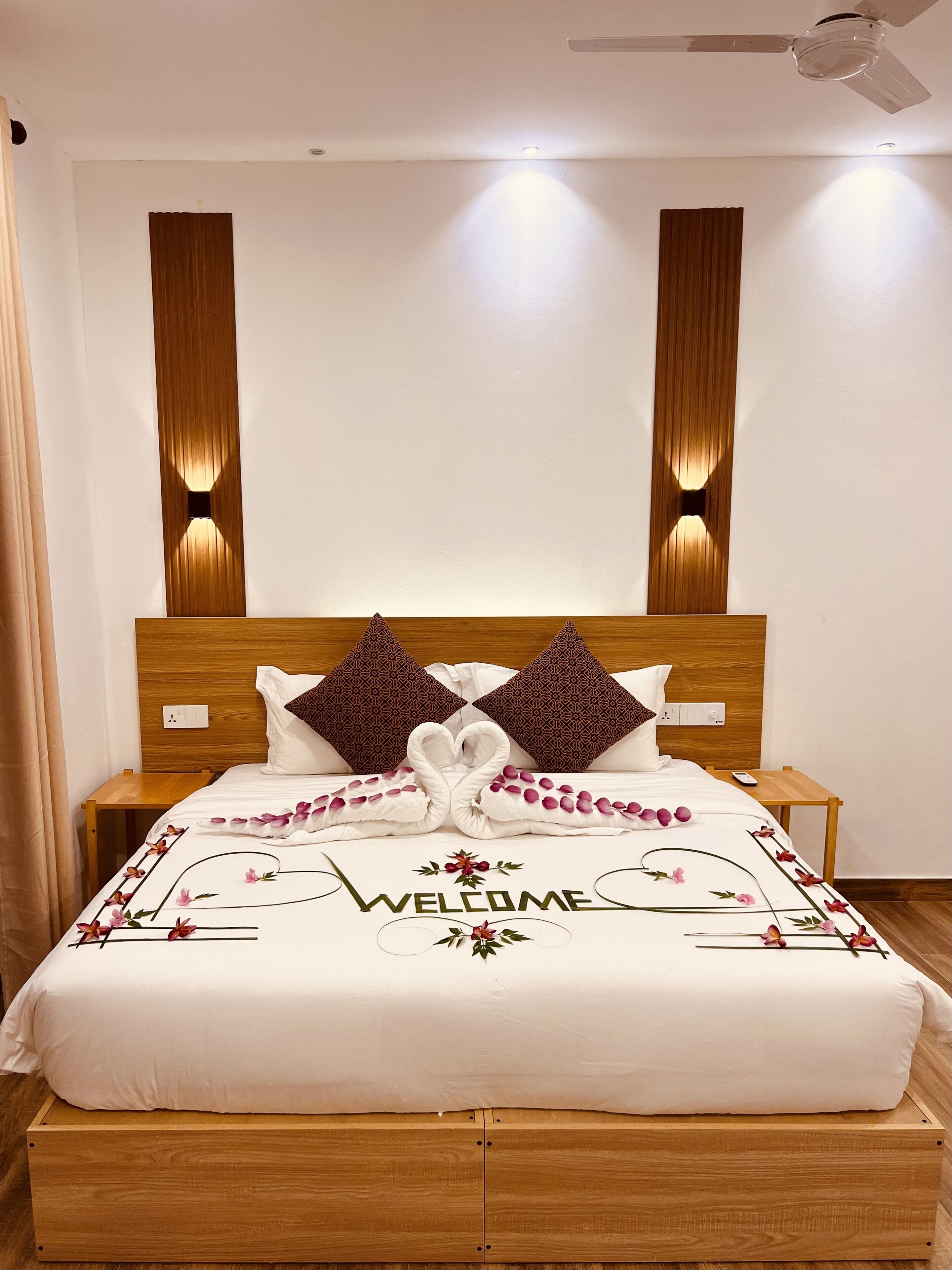 A neatly made bed with a 'WELCOME' sign, wooden headboard, two nightstands, and a ceiling fan.