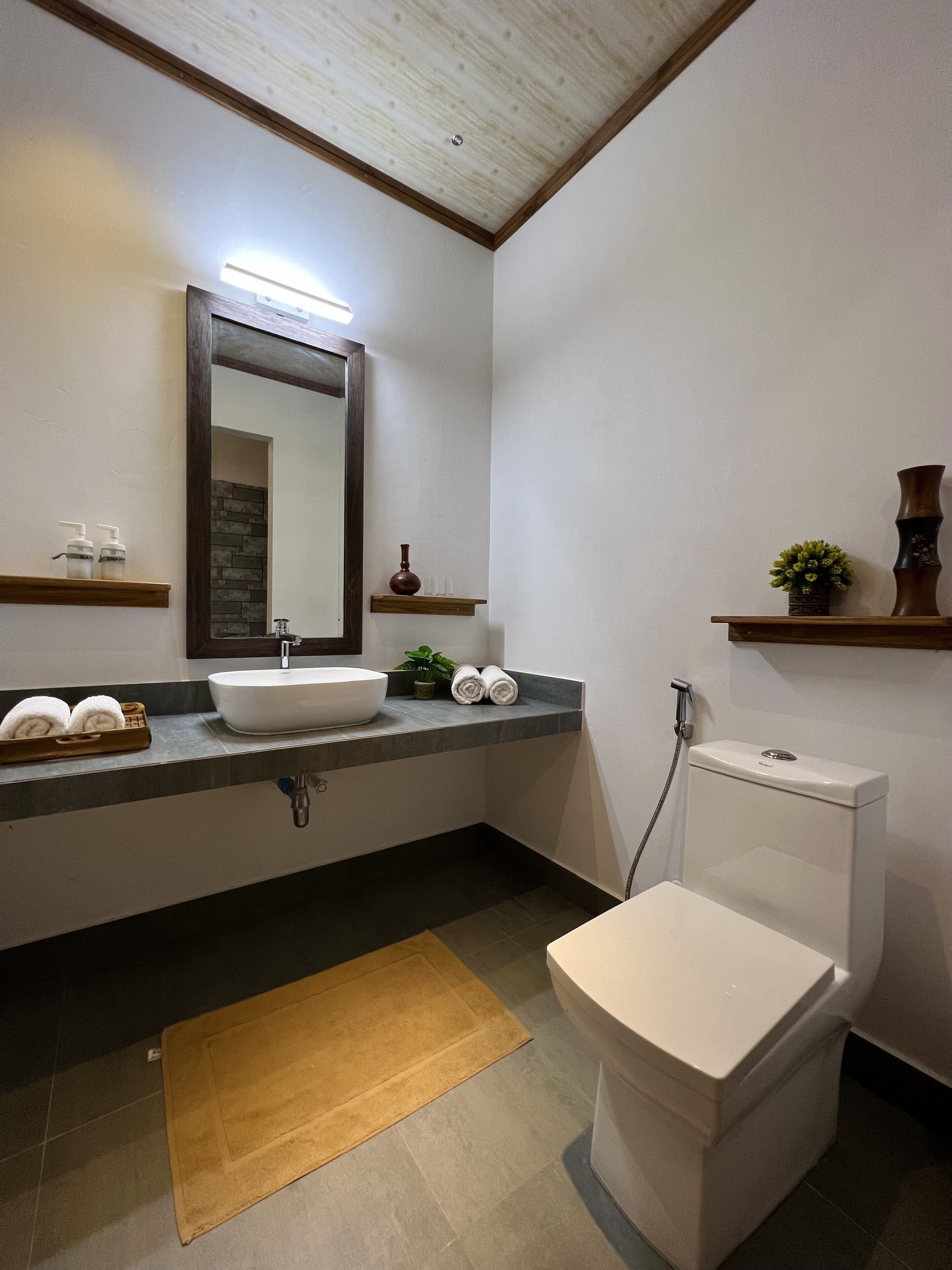 Ein modernes Badezimmer mit Waschbecken, Spiegel und Toilette. Auf der Ablage liegen Handtücher und Pflegeprodukte, und ein kleines Regal mit Dekoration.