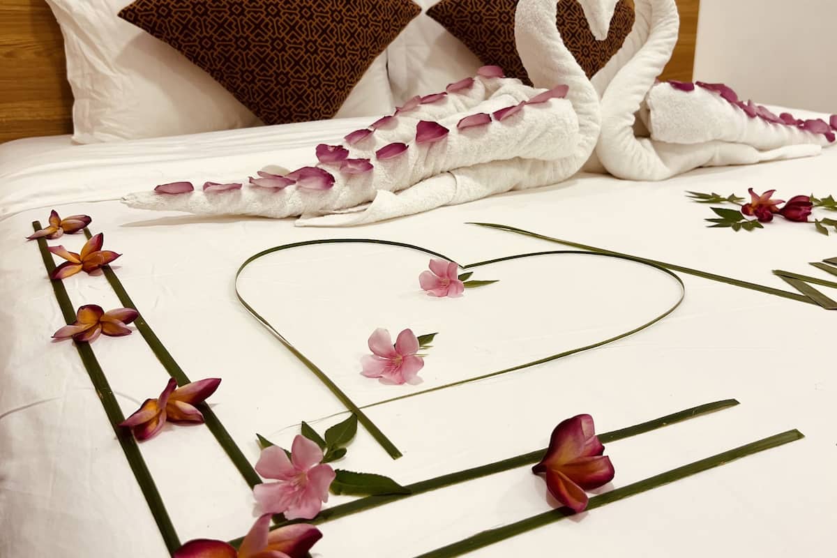 A neatly made bed with a heart-shaped towel arrangement, decorative flowers, and a wooden headboard.