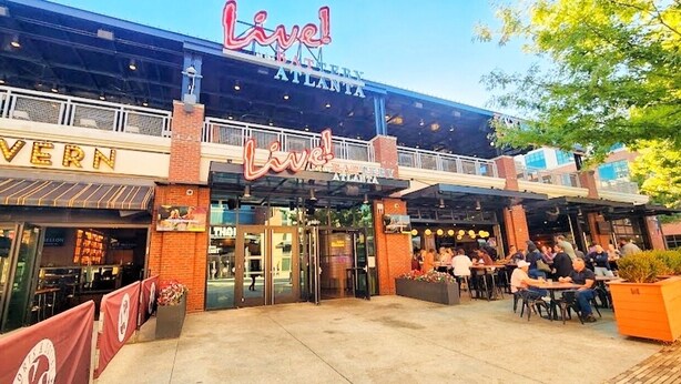 Un área de comedor al aire libre con mesas y sillas, un letrero que dice 'Live! Atlanta', y un edificio con fachada de ladrillo y ventanas de vidrio.