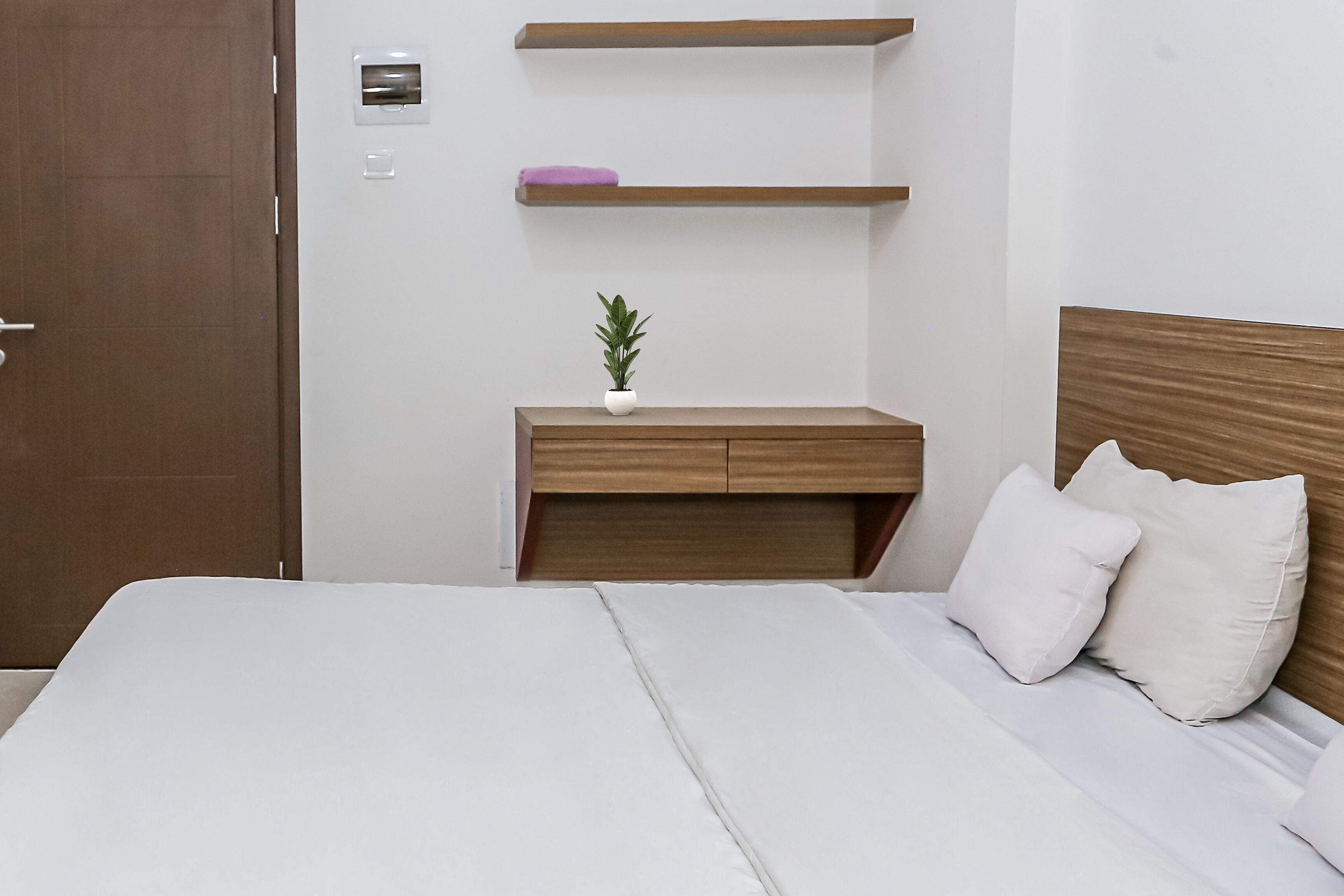 A neatly arranged hotel room with a bed, bedside table, and a small shelf with a plant.