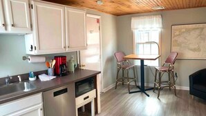 A kitchen with white cabinets, a dishwasher, a coffee maker, and a dining area with two chairs and a table. - Spacious Guest House in Falmouth Village (Falmouth)