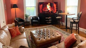 Een woonkamer met een leren bank, een voetenbankje en een flatscreen-tv waarop een haard te zien is.
