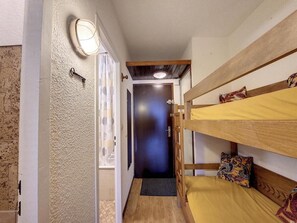 Een smalle gang met twee stapelbedden aan de rechterkant, een deur aan het einde en een lamp aan de linkerkant.