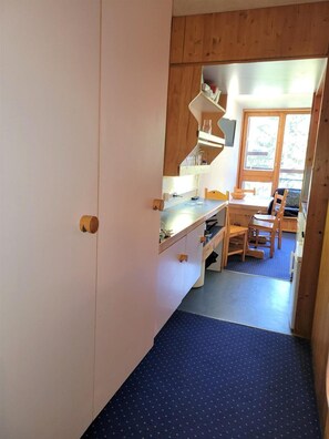 Kompakti keittiö, jossa on puiset kaapit, ruokailutila pöytineen ja tuoleineen sekä näkymä ikkunasta.