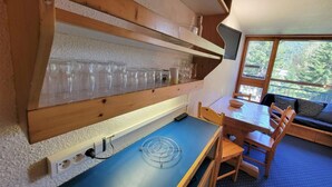 Une pièce chaleureuse avec une table à manger en bois, des chaises, une nappe bleue et une étagère où sont rangés des verres à vin.