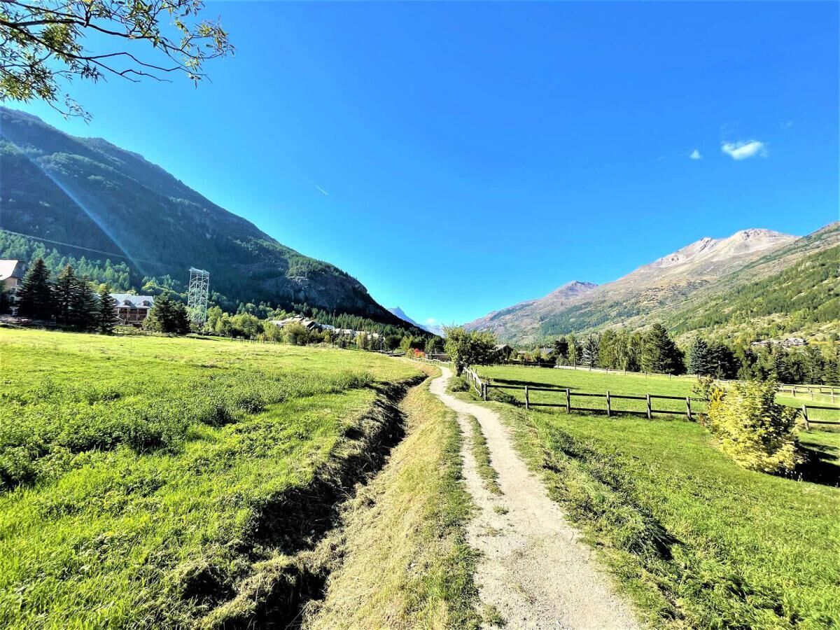 Un paisaje rural con un sendero de tierra, campos verdes y montañas al fondo.