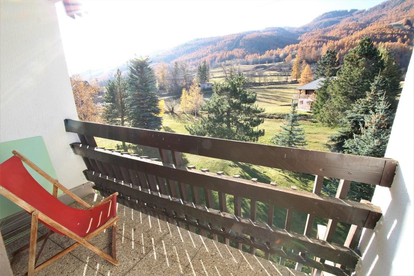 Een balkon met een houten bank en een rode stoel, met uitzicht op een bergachtig landschap met bomen en een klein huis.