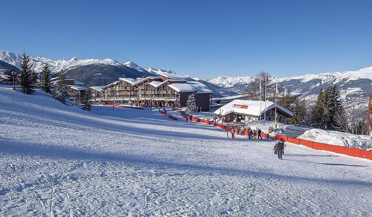 Een skigebied met meerdere berglodge, een skipiste en besneeuwde bergen op de achtergrond.