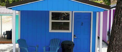 Un cobertizo azul pequeño con una puerta marcada '2', dos sillas azules, un cubo de basura y una mesa de picnic.