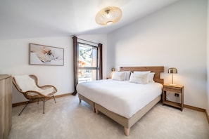 Quarto com cama grande, cadeira de vime, mesa de cabeceira com abajur e janela com cortinas.
