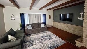 A living room with a grey sofa, a patterned rug, wooden beams on the ceiling, a flat-screen TV mounted on a stone wall, and a potted plant. - Wagner Creek Hideaway (Munising Township)