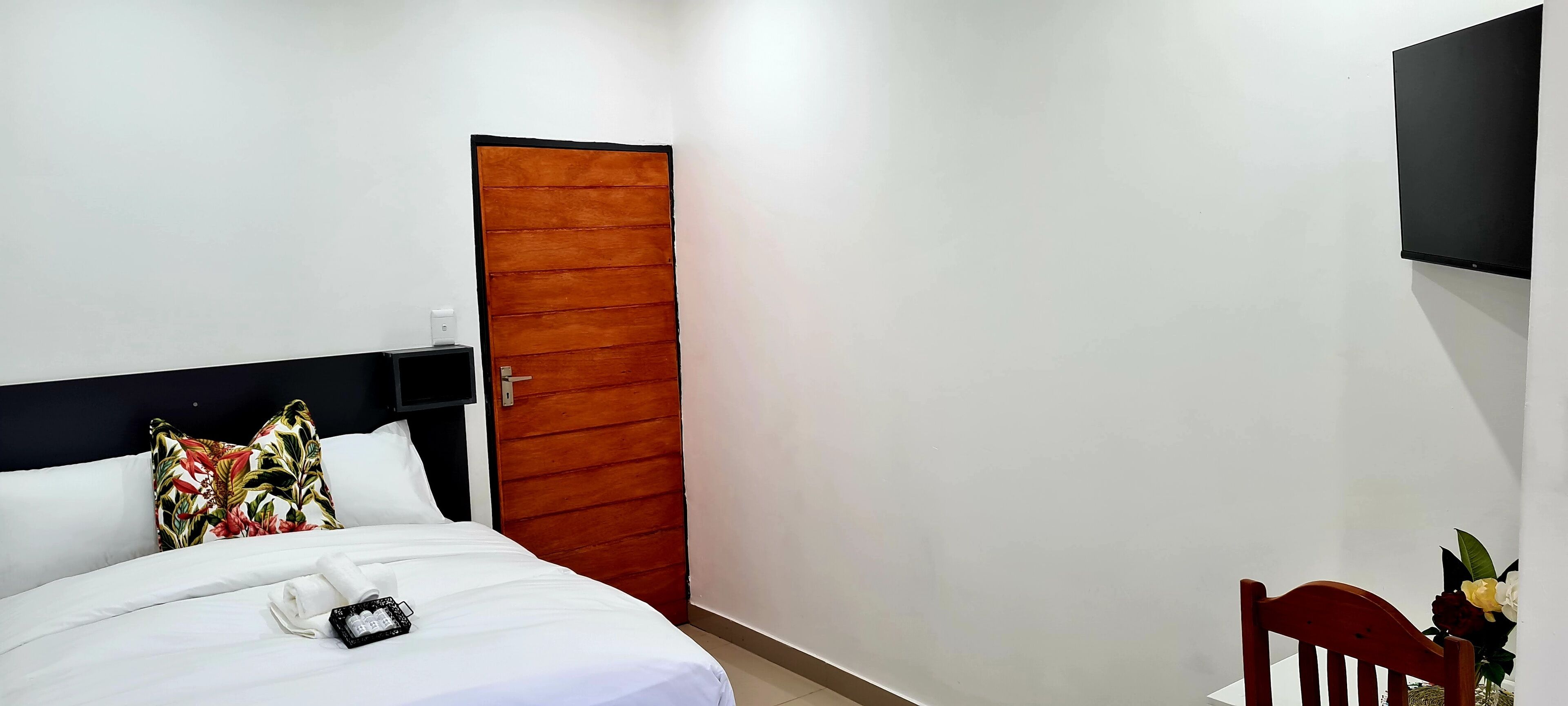 Una camera d'albergo con un letto, una porta in legno, una TV fissata al muro e una sedia con fiori.