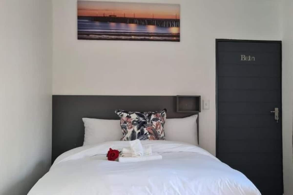 Un lit bien fait, avec du linge de lit blanc, un seul coussin décoratif et une rose rouge. Un tableau encadré est accroché au-dessus du lit, et une porte sombre est visible sur la droite.