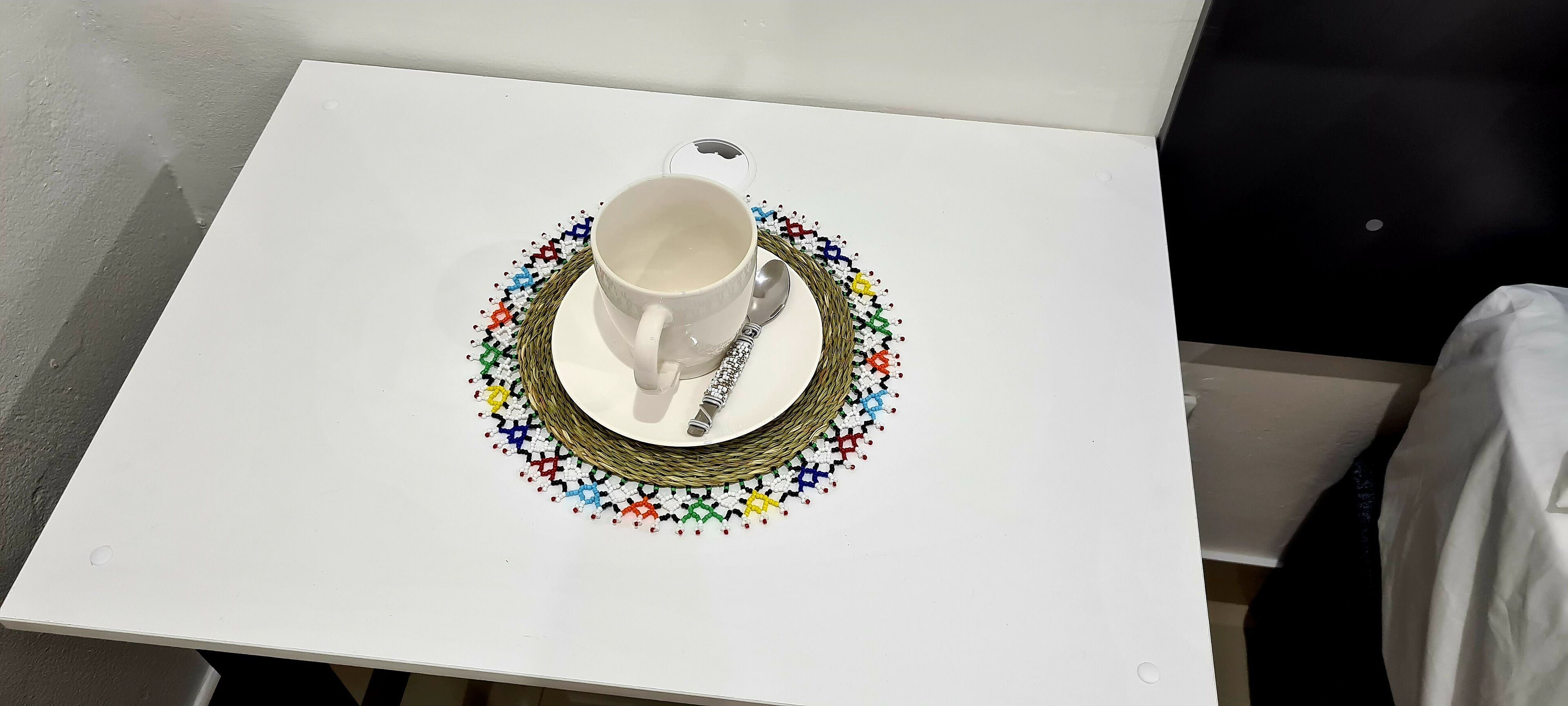 Una mesa blanca con una taza y platillo blancos sobre un posavasos tejido colorido.