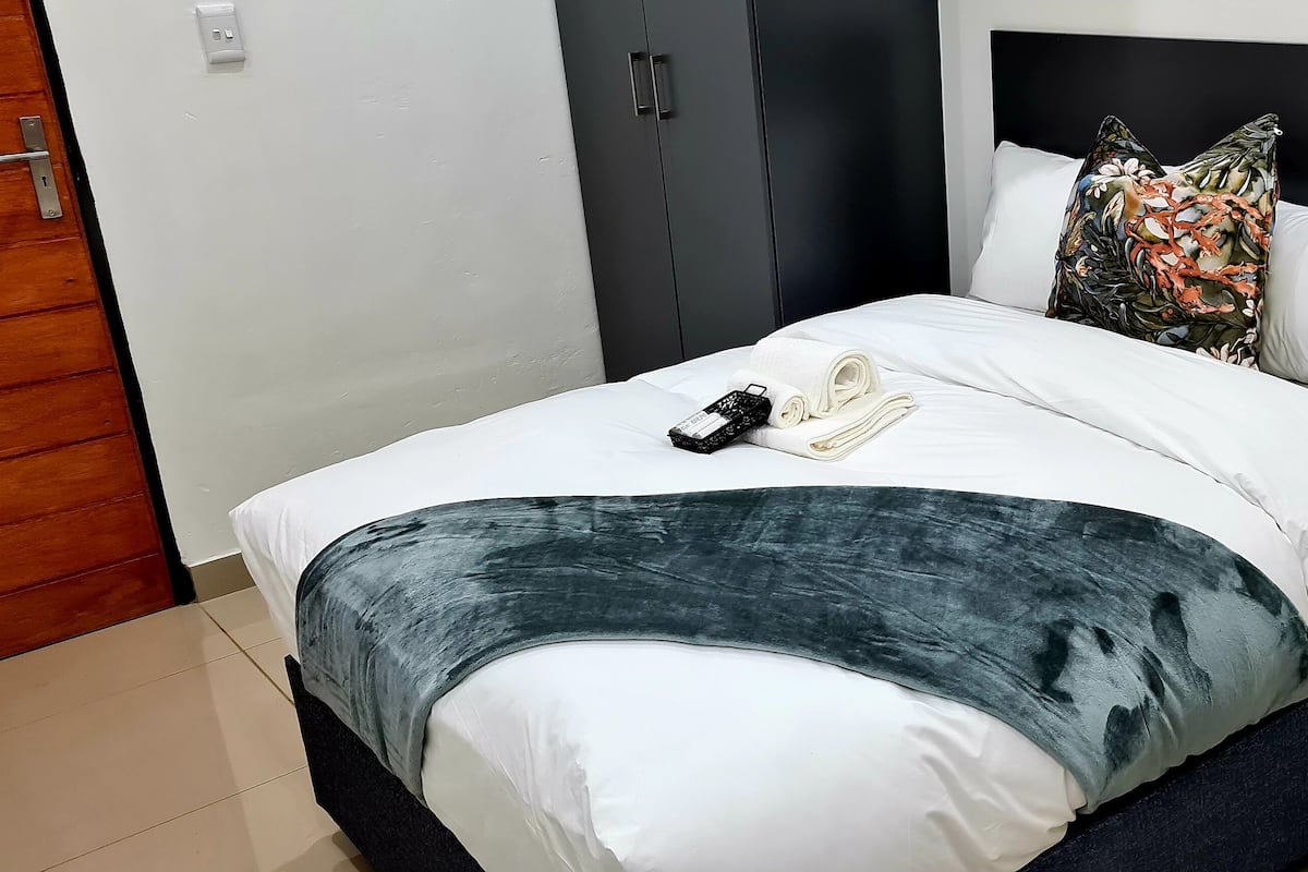 Un lit bien fait, recouvert d’une couverture blanche et bleu foncé, avec une télécommande et une serviette enroulée posées sur le lit.