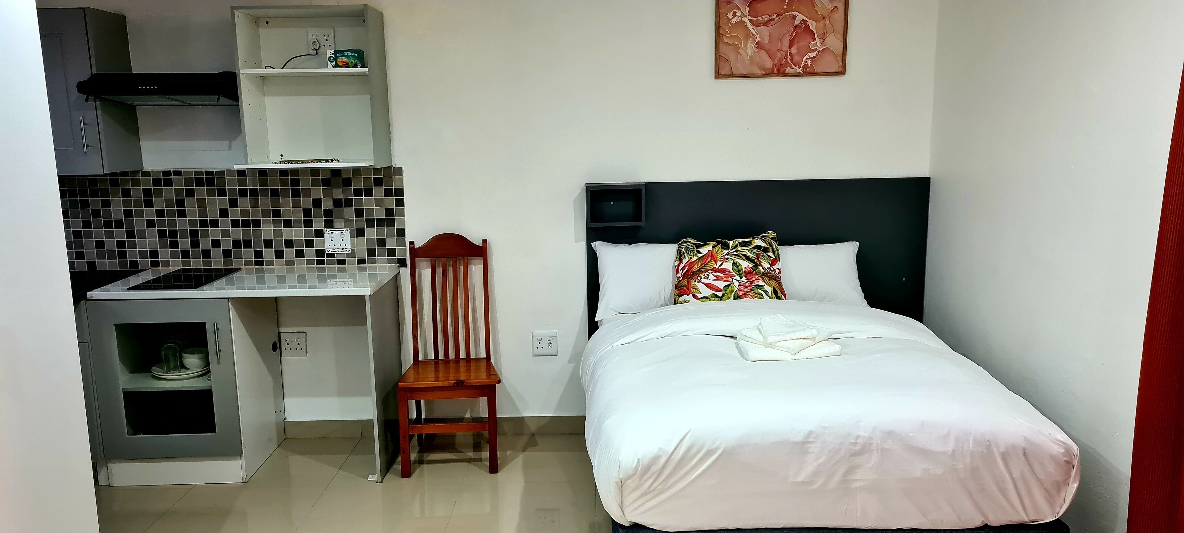 Een kleine kamer met een kitchenette, een bed met een bloemenkussen en een stoel.