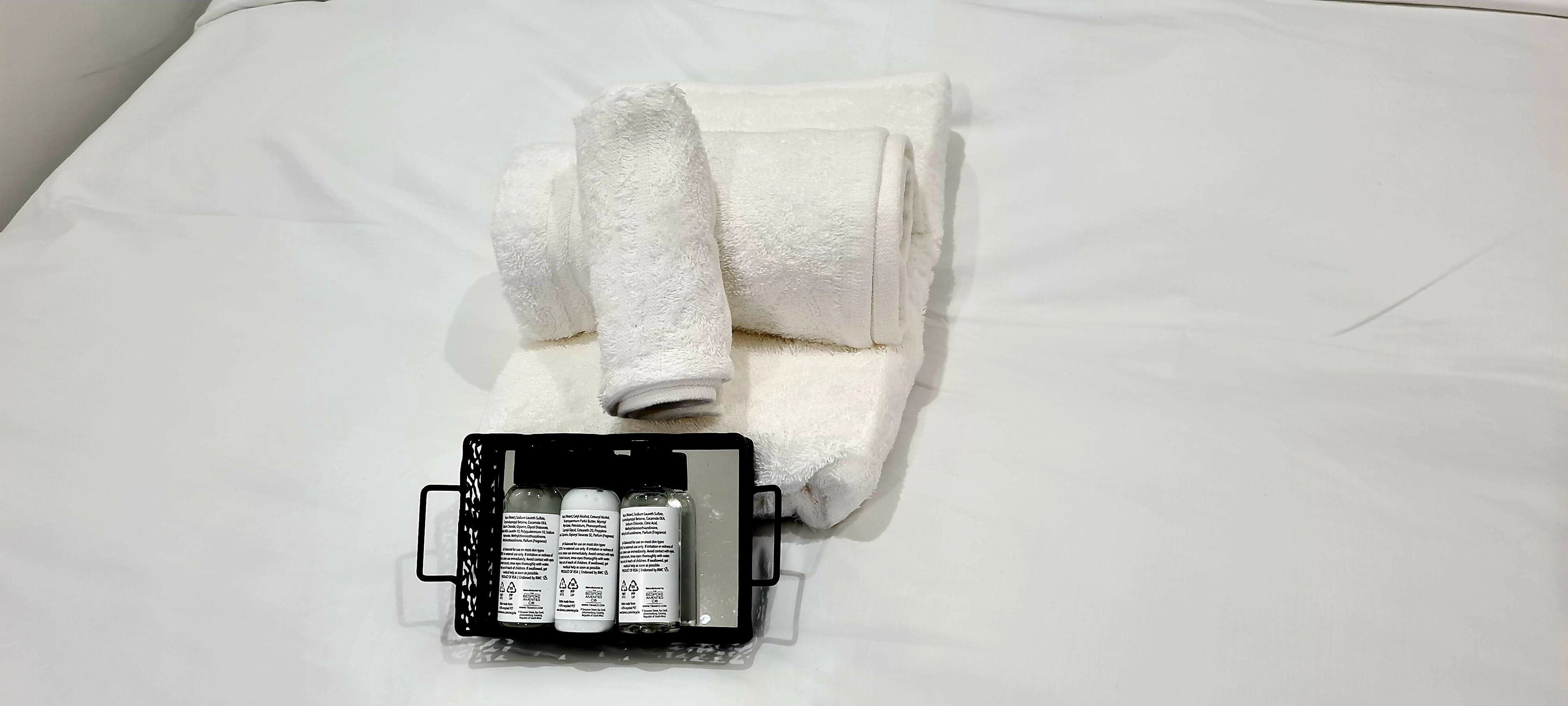 Un llit ben arrenjat amb una tovallola blanca arrotllada i una necesser negre que conté ampolles.