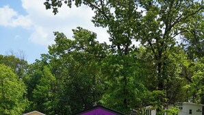 Un petit abri violet avec une porte et une fenêtre, une chaise bleue et une table de pique-nique avec un barbecue et une poubelle devant.